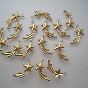 Puede incluir: Un conjunto de colgantes dorados con forma de estrella fugaz. Cada colgante presenta una estrella de cinco puntas con líneas curvas detrás y un pequeño lazo para sujetar a joyas u otras manualidades.