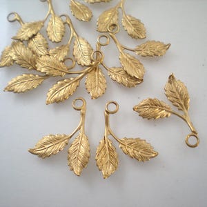Op de afbeelding: Gouden metalen bladcharms met twee lussen om op te hangen. Elke charme heeft een delicate tak met drie bladeren.
