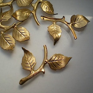 May include: Gold-toned metal leaf sprigs with detailed veining. Each sprig has multiple leaves and a stem. The leaves are arranged in a cluster, with some overlapping.
