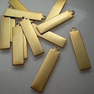 Puede incluir: Un conjunto de etiquetas rectangulares de metal dorado, cada una con un pequeño lazo en un extremo. Las etiquetas tienen un acabado mate y están en blanco, ideales para grabar o estampar. Las etiquetas están dispersas sobre una superficie clara.