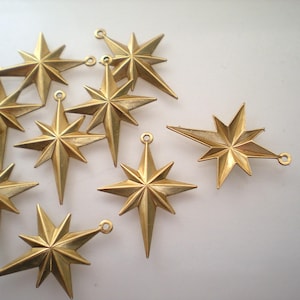 May include: A group of ten gold-colored metal star-shaped charms with a pointed tip. The charms have a hole at the top for hanging.