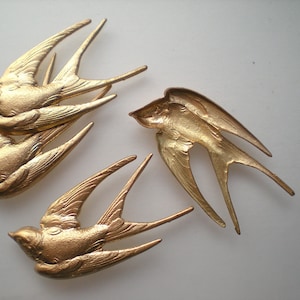 Könnte beinhalten: Mehrere goldfarbene Schwalbenverzierungen sind auf einer weißen Oberfläche angeordnet. Die Vögel sind detailliert mit ausgebreiteten Flügeln und ihren Körpern mit Federn geformt. Die metallische Oberfläche verleiht ihnen ein Vintage- oder Antik-Aussehen, geeignet für Bastel- oder Dekorationsprojekte.