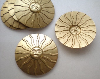 May include: Several gold-colored, circular decorative elements featuring a sun face design with radiating lines. The sun faces have detailed facial features. The elements are arranged in a group, with some overlapping, and are set against a neutral background.