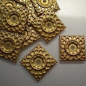 May include: A collection of ornate, square-shaped, gold-colored metal embellishments. Each piece features a central circular design surrounded by a detailed floral pattern. The embellishments have a vintage or antique aesthetic.