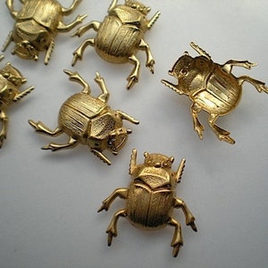 Puede incluir: Cinco figuritas de escarabajo dorado. Los escarabajos están detallados con líneas en relieve y tienen seis patas.