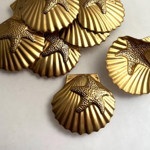 May include: A collection of gold-colored seashell ornaments, each featuring a textured starfish in the center. The shells have a ridged design and a small hole at the top for hanging. These decorative pieces are ideal for coastal-themed decor.