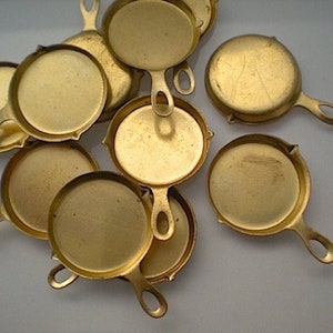May include: A group of ten small, gold-colored, cast iron frying pans with handles. The pans are arranged in a pile on a white surface.