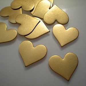 Puede incluir: Una colección de pequeños recortes metálicos en forma de corazón, de color oro cálido. Algunos corazones están enteros, mientras que otros están separados en dos piezas. Los corazones están dispersos sobre una superficie clara.