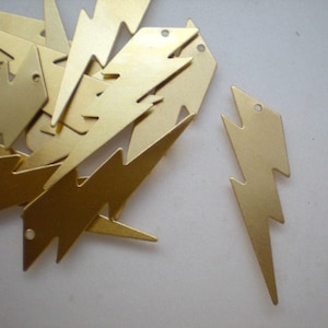 May include: A pile of gold-colored metal lightning bolt charms. The charms are stacked on top of each other and are all the same size and shape. There is one charm that is separated from the pile and is lying flat on the surface.