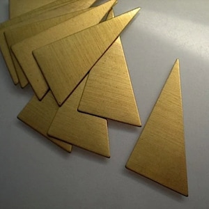 Puede incluir: Una colección de pequeñas piezas metálicas doradas en forma de triángulo. Los triángulos tienen una textura cepillada y están dispuestos sobre una superficie blanca. Las formas son probablemente componentes para joyería o proyectos de artesanía.