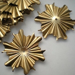 May include: A collection of gold-colored, starburst-shaped metal embellishments. Each piece has a central hole and radiating, ruffled edges. The objects are arranged on a light-colored surface, with some pieces overlapping.
