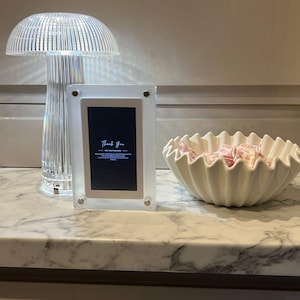 May include: A clear, mushroom-shaped table lamp with a ribbed design stands next to a clear acrylic picture frame. The frame displays the text "Thank You". A white, fluted bowl filled with pink sweets sits nearby.