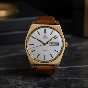 May include: A gold Omega Genève automatic wristwatch with a brown leather strap. The watch face is white with gold hands and markers, and displays the day and date. The watch is set against a dark background.