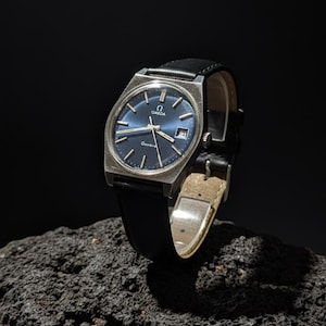 Pode incluir: Um relógio Omega Geneva vintage com caixa quadrada prateada e mostrador azul. O relógio tem uma pulseira de couro preta e é exibido em uma superfície escura e texturizada. O mostrador inclui o logotipo da Omega e a palavra "Geneve".