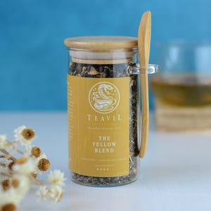 Könnte beinhalten: Ein Glas Teeglas mit der Aufschrift "The Yellow Blend" mit einem HolzLöffel. Das Glas hat einen Holzdeckel und ein gelbes Etikett mit dem Markennamen "TEAVIL" und einem Logo. Im Hintergrund eine Tasse Tee.