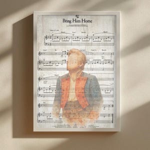 May include: Framed print displaying sheet music with the title "Bring Him Home." The artwork features a watercolour-style illustration of a person in a blue jacket and red vest. The print is set against a light-coloured background.