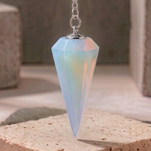 Könnte beinhalten: Ein facettierter, schillernder, opalartiger Pendelanhänger, der an einer silberfarbenen Metallkette hängt. Der Anhänger hat eine spitze, diamantartige Form mit einer glatten, halbtransparenten Oberfläche, die weiches Licht reflektiert. Der Text LIMRA DESIGNS ist sichtbar.