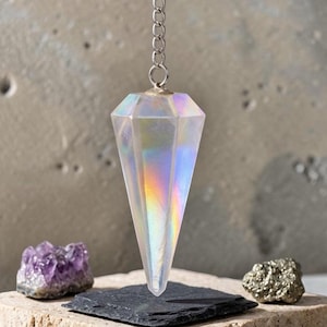 Puede incluir: Un péndulo de cristal transparente, facetado e iridiscente, suspendido de una cadena de metal plateado. El cristal tiene una forma geométrica puntiaguda que refleja colores del arcoíris. Cuelga sobre un cuadrado de pizarra gris oscuro.
