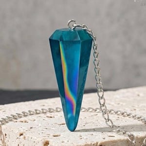 May include: A faceted, teal-colored, cone-shaped pendant with a prominent iridescent rainbow-colored vertical stripe running down its center. The pendant is attached to a silver-toned metal chain that loops around the base of the object.