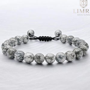 May include: A beaded bracelet featuring round, marbled gray and white stones. The bracelet has a black, adjustable cord closure. The Limra Designs logo is in the upper right corner.
