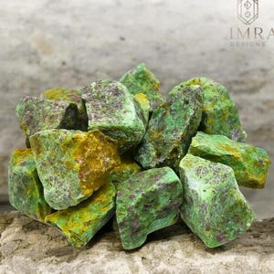 Puede incluir: Un montón de piedras preciosas en bruto verdes y amarillas con motas rojas. Las piedras tienen forma irregular y parecen naturales, con una superficie texturizada. El logotipo "LIMRA DESIGNS" es visible en la esquina superior derecha.