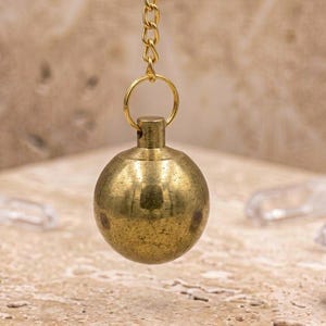 Puede incluir: Un péndulo de metal en tono latón con un peso esférico liso suspendido de una delicada cadena a juego. La esfera tiene un acabado pulido con sutiles variaciones de color, unida a un conector de anillo circular en la parte superior.