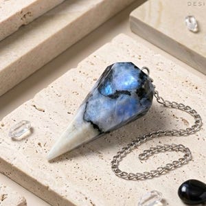 Può includere: Un pendolo in pietra di luna sfaccettato e appuntito con motivi marmorizzati blu e bianchi e inclusioni nere, fissato a una catena in metallo color argento. La pietra presenta una finitura lucida ed è sagomata in una punta geometrica.