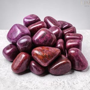 May include: A collection of polished, tumbled gemstones in shades of deep purple and burgundy. The stones have a smooth, glossy finish, with variations in colour and pattern. The image is well-lit, highlighting the natural beauty of the stones. The logo "LIMRA DESIGNS" is in the top right corner.