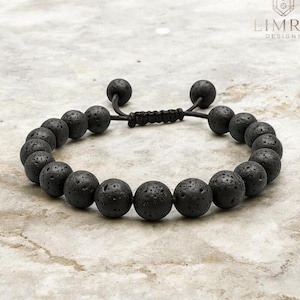 Może przedstawiać: Bransoletka z koralików z czarnego kamienia lawowego z regulowanym sznurkiem. Bransoletka posiada okrągłe, porowate koraliki i zapięcie na przesuwny węzeł. Logo Limra Designs jest widoczne w prawym górnym rogu.
