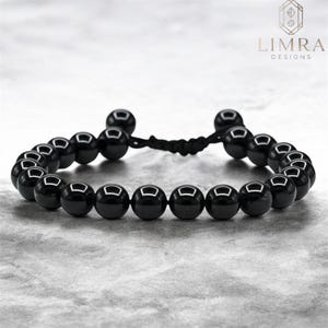 Puede incluir: Una pulsera de cuentas negras con cierre de macramé. La pulsera presenta cuentas negras redondas y pulidas ensartadas en un cordón negro. El logotipo de Limra Designs está en la esquina superior derecha.