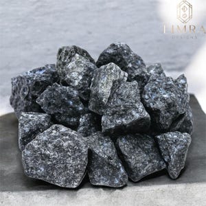 Peut inclure: Un tas de roches gris foncé avec des éclats de gris clair et de bleu. Les roches sont de forme irrégulière et semblent être des pierres naturelles. Le texte "LIMRA DESIGNS" en or est visible.