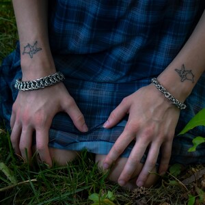 May include: Two silver chainmail bracelets are worn on the wrists. The person is wearing a blue plaid dress and sitting in the grass. Each wrist has a small tattoo of a star.