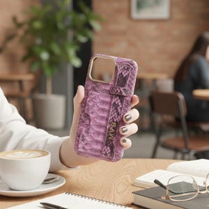 Könnte beinhalten: Eine pinkfarbene Handyhülle mit Schlangenlederstruktur und einem Riemen. Die Hülle hat einen goldfarbenen Kameraausschnitt und die Initialen "MVB" sind auf dem Riemen zu sehen. Die Hülle wird vor einem verschwommenen Café-Hintergrund gehalten.