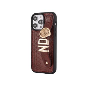May include: A brown textured phone case with a black border. The case features a gold-colored ring holder with a geometric pattern and the gold letters "ND". The case has a strap with a gold stud.