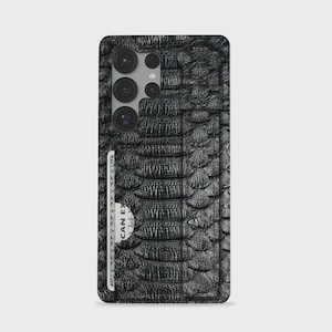 May include: Black phone case with a textured, snake-skin pattern. The case has a built-in card holder with a white card partially visible. The phone's camera lenses are visible at the top. The case is designed to fit a smartphone.