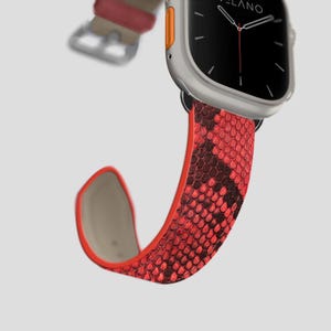 May include: A silver smartwatch with a black screen and a red snakeskin-patterned strap. The strap is red with dark brown accents. The watch has an orange button on the side.