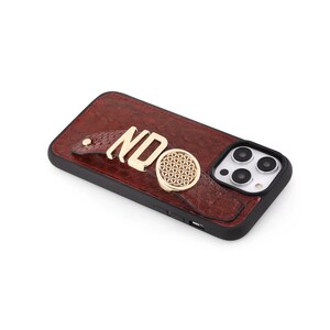 May include: A brown leather phone case with a gold-colored strap and a gold-colored ring holder. The case has the letters "NDO" in gold. The phone case has a black border and a camera cutout.