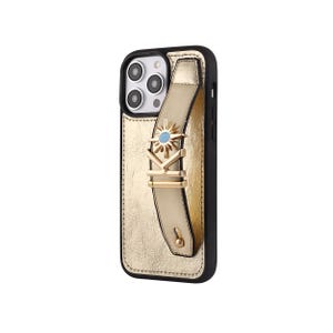May include: Gold-colored phone case with a black border. The case features a gold strap with a decorative sunburst and the letters "K" and "I" in gold. The strap has a small blue gem in the center of the sunburst.