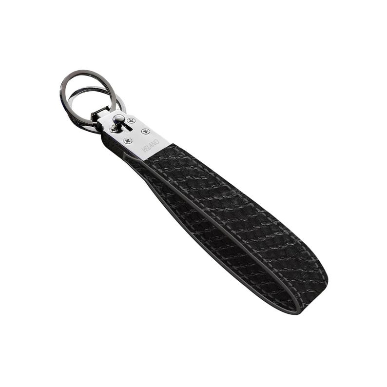 May include: Black leather keychain wrist strap with a woven pattern. The strap is attached to a silver metal clasp and a dark metal key ring. The word "VELANO" is printed on the clasp.