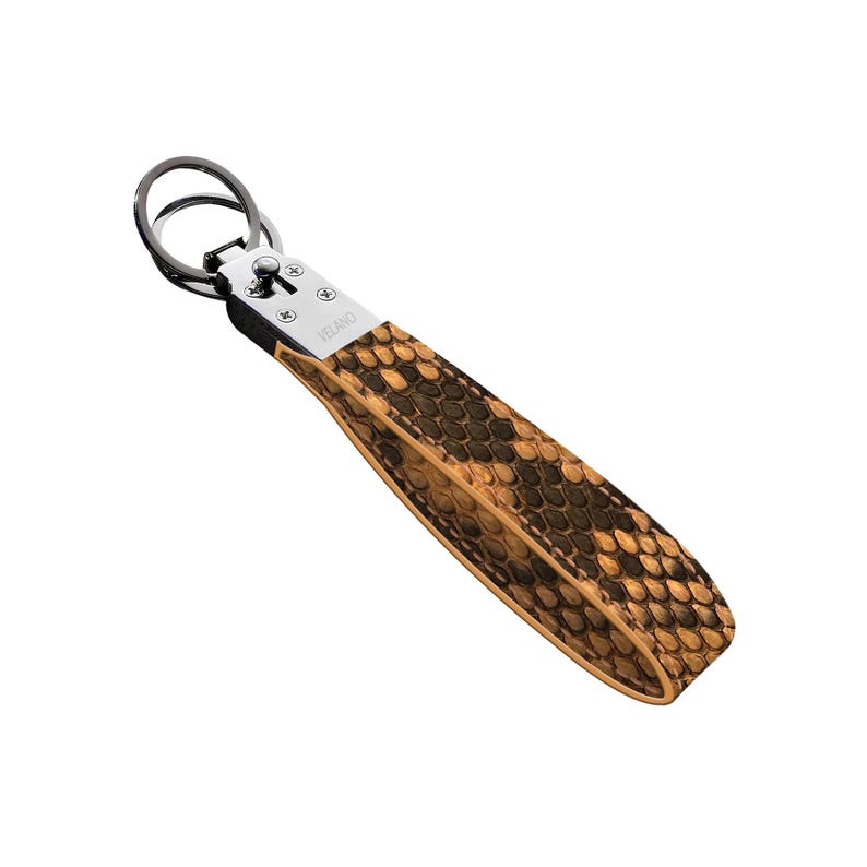 May include: A leather keychain with a snakeskin pattern in shades of brown and tan. The keychain has a silver-colored metal ring and a small metal plate with the word "VELANO" engraved on it.