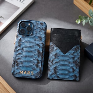 Op de afbeelding: Een blauw en zwart telefoonhoesje met slangenleertextuur en de naam MARS in goud reliëf, gecombineerd met een bijpassende kaarthouder met slangenleertextuur, een zwart lederen bovendeel en een gouden ritstrekker.