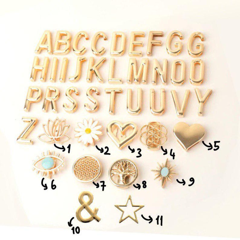 May include: Gold-colored metal alphabet letters and charms. The charms include a lotus flower, daisy, heart, and star. The letters and charms are arranged on a white surface.