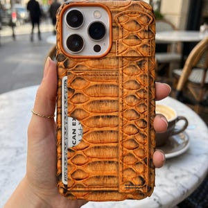 Könnte beinhalten: Eine Smartphone-Hülle mit strukturiertem orange-schwarzem Schlangenledermuster, ausgestattet mit einem integrierten Kartenfach, das eine Karte mit dem sichtbaren Text CAN EX enthält, sowie einem vertikalen Halteband um die Dreifachkamera.