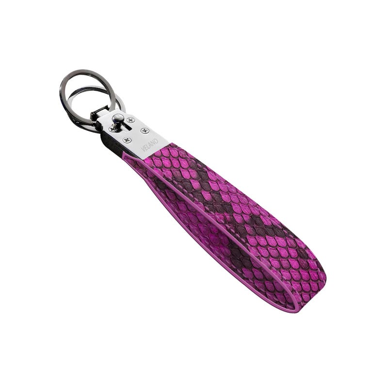 May include: A key chain wrist strap with a vibrant pink and black snakeskin pattern. The strap is attached to a silver-tone metal ring and hardware. The word "VELANO" is printed on the strap.