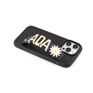 May include: Black textured phone case with gold lettering spelling "ADA" and a white daisy with a yellow centre. The case has a camera cutout and is designed for a smartphone.