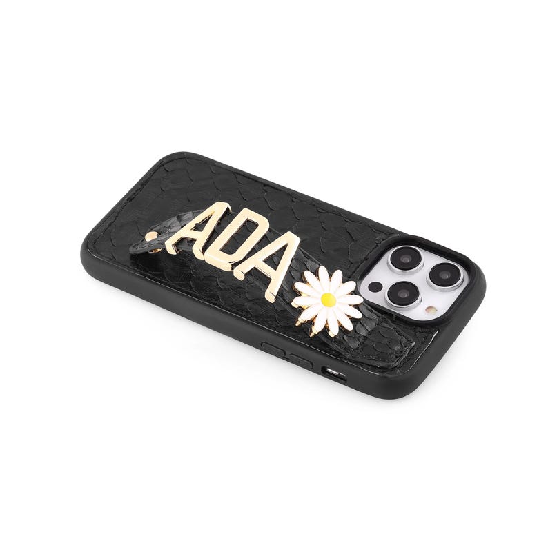 May include: Black textured phone case with gold lettering spelling "ADA" and a white daisy with a yellow centre. The case has a camera cutout and is designed for a smartphone.