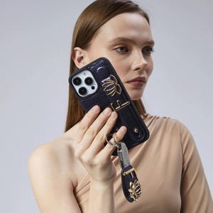 May include: A dark blue phone case with a gold lotus flower design and a matching wrist strap. The case has a textured finish and a gold metal accent. A silver keychain with the brand name is attached to the wrist strap.