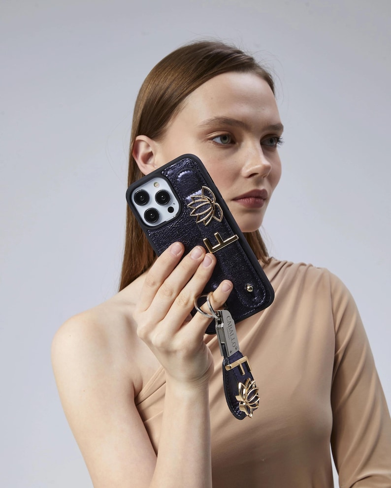May include: A dark blue phone case with a gold lotus flower design and a matching wrist strap. The case has a textured finish and a gold metal accent. A silver keychain with the brand name is attached to the wrist strap.