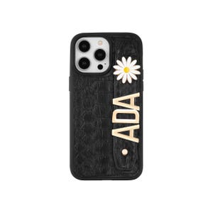 Könnte beinhalten: Schwarze Handyhülle mit strukturiertem Muster, einer weißen und gelben Gänseblümchenblüte und dem Namen "ADA" in goldfarbenen Buchstaben. Die Hülle hat einen Kameraausschnitt und eine Schlaufe für eine einfache Handhabung.