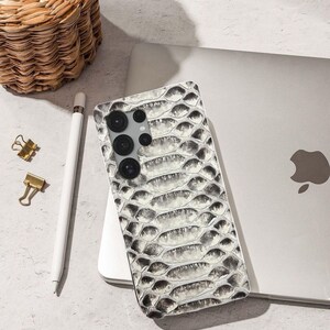 Puede incluir: Una funda de teléfono inteligente con un patrón de piel de serpiente en tonos grises y blancos. La funda está sobre un portátil, junto a un lápiz óptico blanco, clips dorados y una cesta de mimbre.